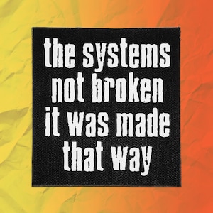 May include: A square black fabric patch featuring white text that reads, the systems not broken it was made that way. The lettering is in a bold, sans-serif font with a slightly distressed, textured appearance against the dark background.