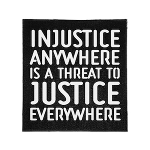 Peut inclure: Un écusson carré en tissu noir avec le texte blanc suivant : INJUSTICE ANYWHERE IS A THREAT TO JUSTICE EVERYWHERE. Les lettres sont dans une police grasse sans empattement, et l'écusson présente une texture tissée visible sur le fond noir.