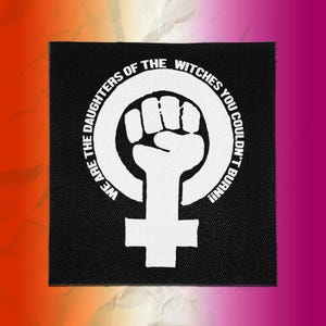 May include: A square black fabric patch featuring a white screen-printed design of a raised fist inside a female gender symbol. The text "WE ARE THE DAUGHTERS OF THE WITCHES YOU COULDN'T BURN!!" is printed in a circular arc around the central graphic.