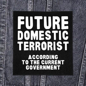 Pode incluir: Patch quadrado preto com texto branco: "FUTURE DOMESTIC TERRORIST" acima de "ACCORDING TO THE CURRENT GOVERNMENT". O patch está em uma jaqueta jeans escura.