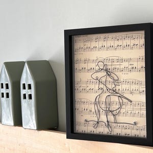 Könnte beinhalten: Gerahmtes Kunstwerk mit einer schwarzen Drahtkontur eines Cellisten vor einem Hintergrund aus Notenblättern. Zwei grüne Keramik-Hausdekorationen befinden sich links.