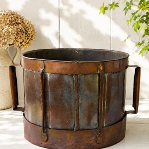 May include: A cylindrical copper planter with a weathered, oxidised patina featuring vertical metal bands and two side handles. The vessel has a wide open top and a dark, aged finish with visible green and brown tones throughout the metal surface.