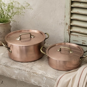 May include: Two round copper pots with matching lids and brass handles. The larger pot is taller, while the smaller pot is shallower. Both feature polished metallic surfaces and sturdy side handles, showcasing a classic design for kitchen cookware.