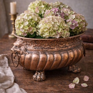 May include: A copper-toned decorative planter filled with green and pink hydrangeas. The planter features lion head handles and claw feet. The arrangement sits on a dark wooden surface, with a candle and fabric in the background.