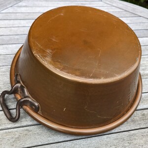 Op de afbeelding: Een koperen pot met twee ijzeren handvatten. De pot heeft een versleten, verweerde uitstraling en staat op een houten oppervlak.