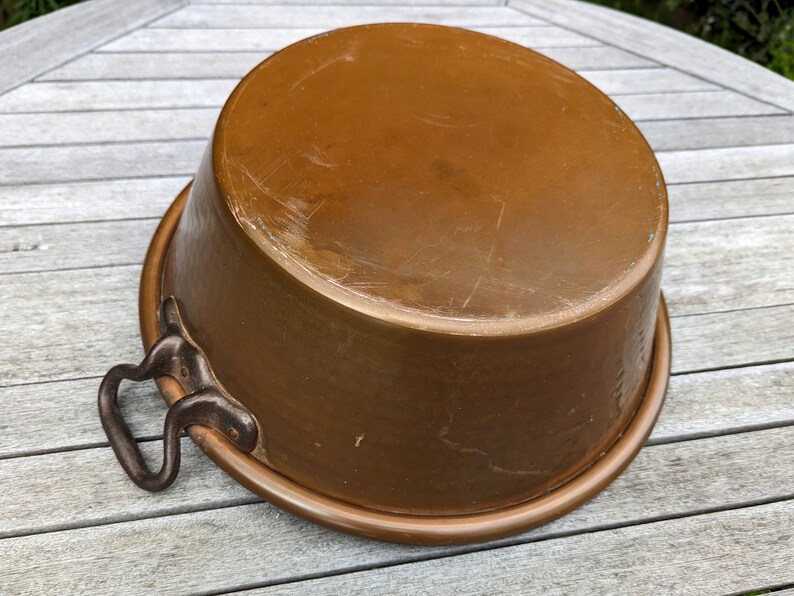 Op de afbeelding: Een koperen pot met twee ijzeren handvatten. De pot heeft een versleten, verweerde uitstraling en staat op een houten oppervlak.