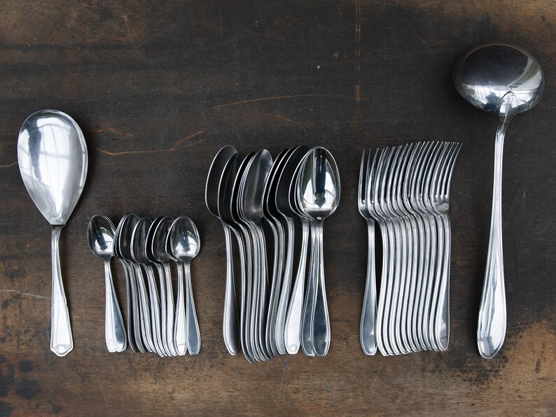 May include: A set of silverware, including a serving spoon, a ladle, forks, and spoons, all laid out on a dark wooden surface.