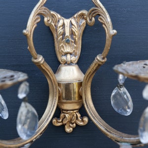 K&ouml;nnte beinhalten: Eine goldfarbene Wandleuchte mit verzierten Details und Kristalltropfen. Die Leuchte hat ein dekoratives Rankendesign und ist an einer Wand montiert.