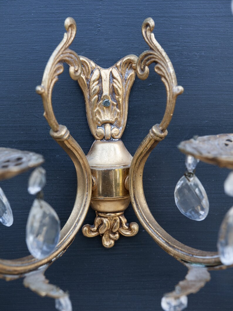 K&ouml;nnte beinhalten: Eine goldfarbene Wandleuchte mit verzierten Details und Kristalltropfen. Die Leuchte hat ein dekoratives Rankendesign und ist an einer Wand montiert.