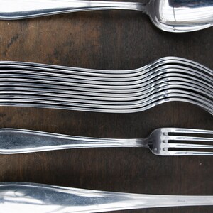 May include: A set of silverware, including a spoon, fork, and a stack of forks. The silverware is made of polished stainless steel and is arranged on a wooden surface.