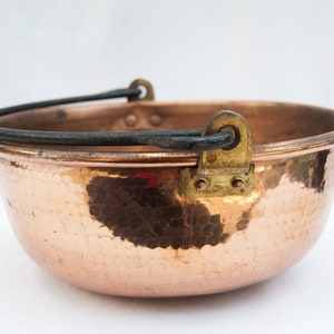 May include: A copper cauldron with a hammered texture and a black metal handle. The cauldron has a rounded shape and is designed with brass hardware for the handle attachment. The interior of the cauldron is a shiny copper color.