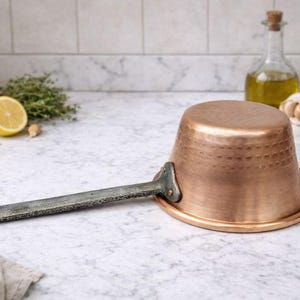 May include: A copper saucepan with a long, gray handle and a hammered texture. The pan is resting on a marble surface, with a lemon, herbs, and olive oil in the background. A classic kitchenware item.