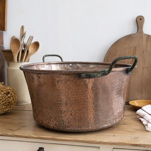 May include: A large, round, hammered copper basin featuring a wide opening and two dark, weathered metal handles attached to the sides. The exterior surface displays a textured, dimpled finish with natural colour variations in the copper metal.