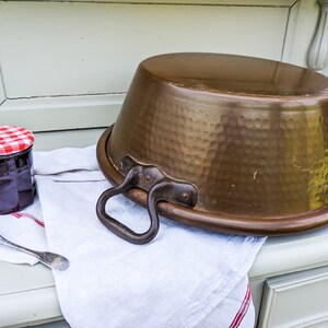 Op de afbeelding: Een grote, gehamerde koperen pot met een handvat staat op een witte doek met een rode en witte geruite rand. Op de achtergrond staat een pot jam met een rood en wit geruit deksel.