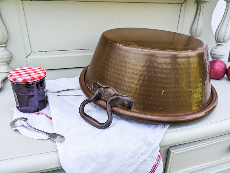Op de afbeelding: Een grote, gehamerde koperen pot met een handvat staat op een witte doek met een rode en witte geruite rand. Op de achtergrond staat een pot jam met een rood en wit geruit deksel.