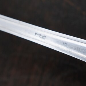 May include: Close-up of a silver utensil with the words "Lumen Inox" engraved on the handle.