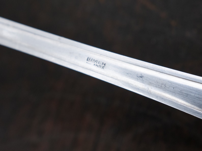 May include: Close-up of a silver utensil with the words "Lumen Inox" engraved on the handle.