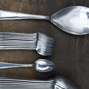 May include: A close-up of a set of silver spoons, including a large serving spoon, a smaller spoon, and a stack of 12 spoons. The spoons are arranged vertically on a dark brown wooden surface.