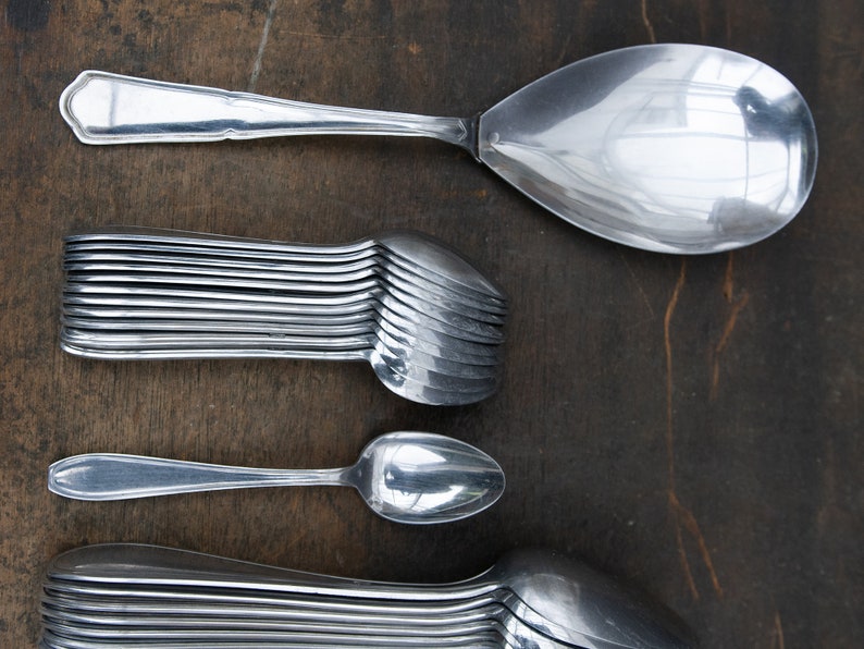 May include: A close-up of a set of silver spoons, including a large serving spoon, a smaller spoon, and a stack of 12 spoons. The spoons are arranged vertically on a dark brown wooden surface.