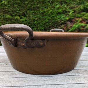 Op de afbeelding: Een grote, gehamerde koperen kom met een enkele, zwarte ijzeren handgreep. De kom staat op een houten oppervlak.