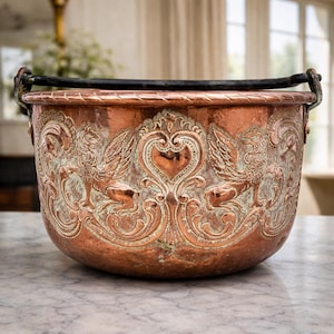 Puede incluir: Un gran caldero de cobre antiguo con un asa negra. El caldero presenta tallas en relieve ornamentadas de criaturas míticas y diseños en forma de corazón. El cobre tiene una rica pátina envejecida con toques verdes.