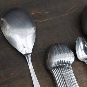 May include: A set of silver spoons, including a large serving spoon, a smaller spoon, and a bundle of spoons. The spoons are arranged on a dark brown wooden surface.