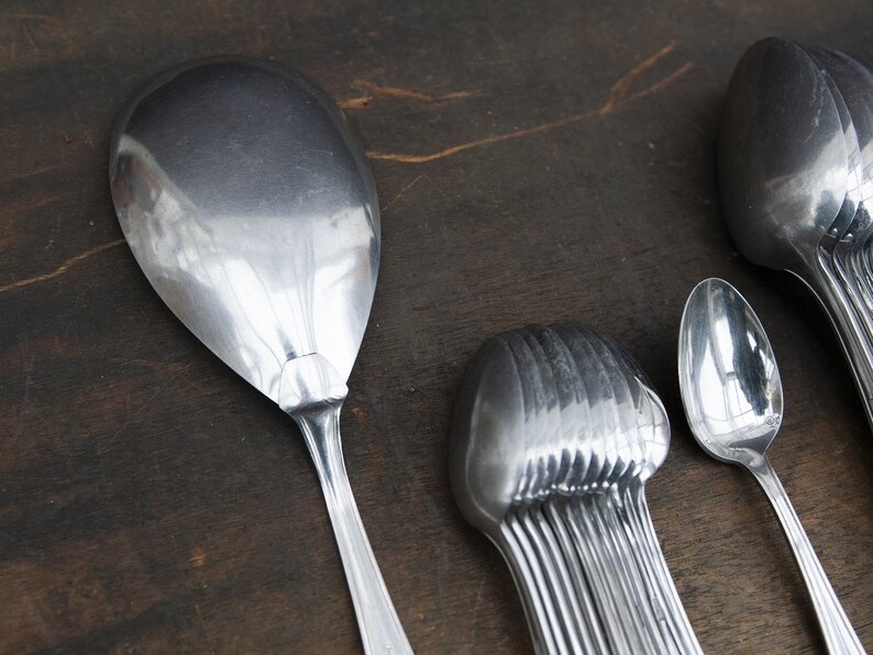 May include: A set of silver spoons, including a large serving spoon, a smaller spoon, and a bundle of spoons. The spoons are arranged on a dark brown wooden surface.