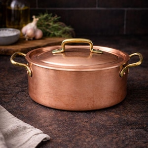 Puede incluir: Una olla de cocina de color cobre con tapa y asas doradas. La olla es redonda y se asienta sobre una superficie marrón oscuro. La tapa tiene un solo asa y la olla tiene dos asas laterales.