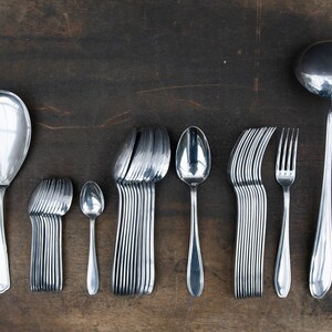 May include: A set of silverware, including a large serving spoon, a set of smaller spoons, a set of forks, and a ladle. The silverware is arranged in a row on a wooden surface.