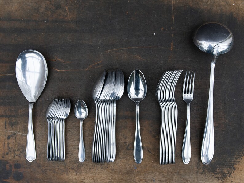 May include: A set of silverware, including a large serving spoon, a set of smaller spoons, a set of forks, and a ladle. The silverware is arranged in a row on a wooden surface.