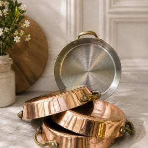 May include: A stack of three round copper pans with polished metallic interiors and brass handles. The pans feature a warm, reflective exterior finish and are arranged with one pan standing upright and two pans nested horizontally below it.