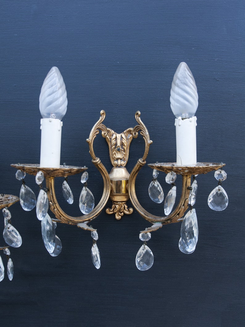 K&ouml;nnte beinhalten: Eine goldfarbene Wandleuchte mit zwei mattierten Gl&uuml;hbirnen und Kristalltropfen. Die Leuchte hat ein dekoratives, geschwungenes Design.