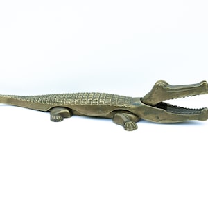 May include: A brass alligator nutcracker with an open mouth, ready to crack nuts. The nutcracker is on a white surface.