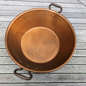 Op de afbeelding: Een grote koperen pot met een gehamerde textuur en twee ijzeren handvatten. De pot is leeg en staat op een houten oppervlak.