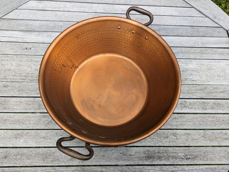 Op de afbeelding: Een grote koperen pot met een gehamerde textuur en twee ijzeren handvatten. De pot is leeg en staat op een houten oppervlak.