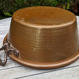 Op de afbeelding: Een grote, gehamerde koperen pot met een enkele ijzeren handgreep. De pot heeft een ronde bodem en een brede, ondiepe opening.