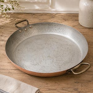May include: A round, shallow copper pan with a hammered silver-toned interior lining. The pan features two curved, dark metallic handles attached to the sides with three visible rivets each. The exterior rim shows the warm, reddish-brown copper finish.