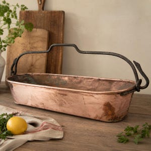 Op de afbeelding: Een langwerpig, rechthoekig koperen bekken met een verweerde, metalen afwerking en een donker, getextureerd ijzeren handvat dat aan elke kant is bevestigd met decoratieve, haakvormige metalen lussen. De binnenkant vertoont sporen van oxidatie.