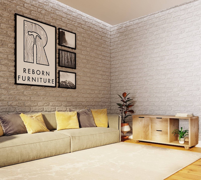 May include: A light beige sofa with yellow and gray throw pillows sits in front of a white brick wall. Three black framed pictures hang on the wall, one of which features the text "REBORN FURNITURE". A wooden cabinet with two drawers and an open shelf sits on the right side of the image. A potted plant sits on the shelf.