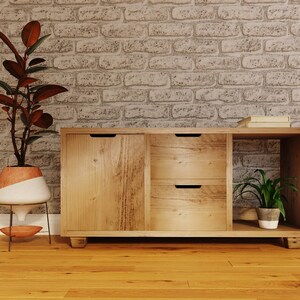 May include: A wooden cabinet with two drawers and a door. The cabinet has a light wood finish and is sitting on a wooden floor. There is a plant in a pot on the top shelf of the cabinet.