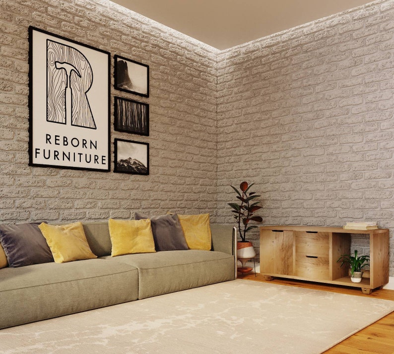 May include: A light brown wooden cabinet with two drawers and a shelf. The cabinet has a rustic look and is sitting on a light brown wooden floor. The cabinet is in front of a white brick wall with a gray couch and yellow pillows. There are three black framed pictures on the wall with the text "REBORN FURNITURE" on the largest picture.