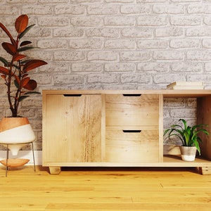 May include: A light wood cabinet with two drawers and a door. The cabinet has a shelf on the right side. A potted plant is on the shelf. The cabinet is in front of a white brick wall.