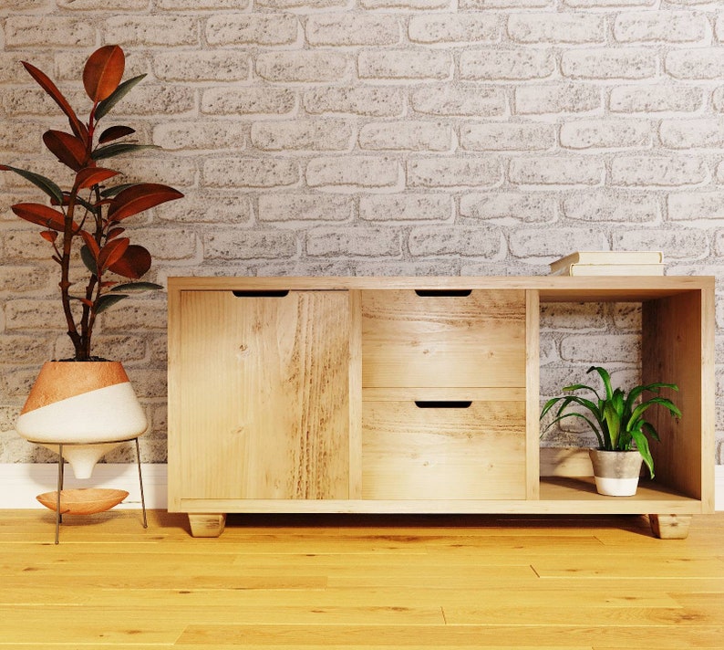 May include: A light wood cabinet with two drawers and a door. The cabinet has a shelf on the right side. A potted plant is on the shelf. The cabinet is in front of a white brick wall.