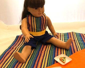 Puede incluir: Una muñeca con una camiseta a rayas y pantalones cortos azules está sentada sobre una manta a rayas colorida. Dos pequeños platos blancos contienen porciones de pizza. Un lazo amarillo adorna el cabello de la muñeca. Tema de picnic.