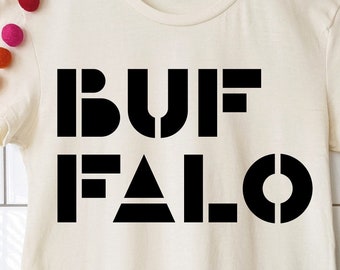 May include: Cream-colored t-shirt with the word "BUFFALO" printed in bold, black, stencil-style letters. The shirt is hanging on a wooden hanger. A string of colorful felt balls decorates the upper left corner.