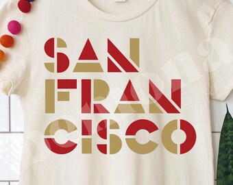 Puede incluir: Una camiseta beige con un diseño gráfico de las palabras "SAN FRANCISCO" en letras de bloque rojas y doradas.