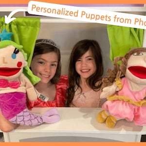 May include: Two personalized puppets made from photos. The puppets are a mermaid with green hair and a pink tail and a princess with brown hair and a pink dress. The puppets are sitting on a white shelf with a green curtain behind them.