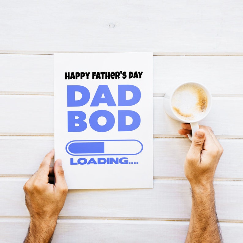 Pode incluir: Um cart&atilde;o branco com o texto "Happy Father's Day Dad Bod Loading..." em letras pretas e azuis. Uma barra de progresso azul est&aacute; parcialmente preenchida. Uma m&atilde;o segura o cart&atilde;o. Uma x&iacute;cara branca de caf&eacute; com um l&iacute;quido marrom est&aacute; sobre uma superf&iacute;cie de madeira branca.