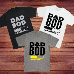 Pode incluir: Tr&ecirc;s camisetas com o texto "Dad Bod Loading..." impresso nelas. As camisetas s&atilde;o preta, branca e cinza. A barra de progresso em cada camiseta est&aacute; em um est&aacute;gio diferente de conclus&atilde;o.
