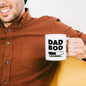 Pode incluir: Caneca de cer&acirc;mica branca com um gr&aacute;fico preto que diz "Dad Bod Loading..." com uma barra de progresso parcialmente preenchida.